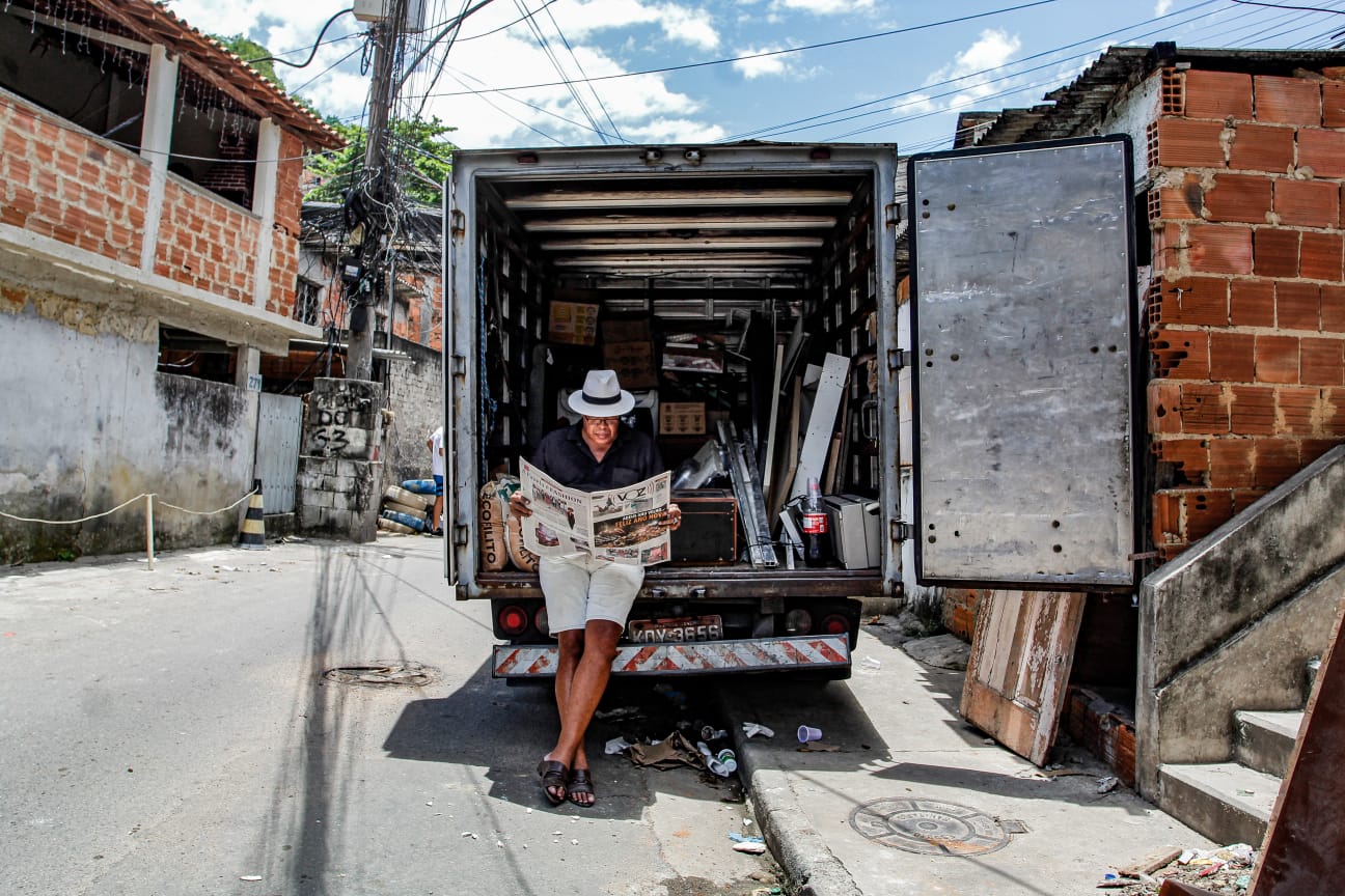 Um homem de chapéu branco lê um jornal do Voz das Comunidades encostado na traseira aberta de um caminhão carregado, estacionado em uma rua estreita do Complexo do Alemão. Foto: Uendell Vinicius