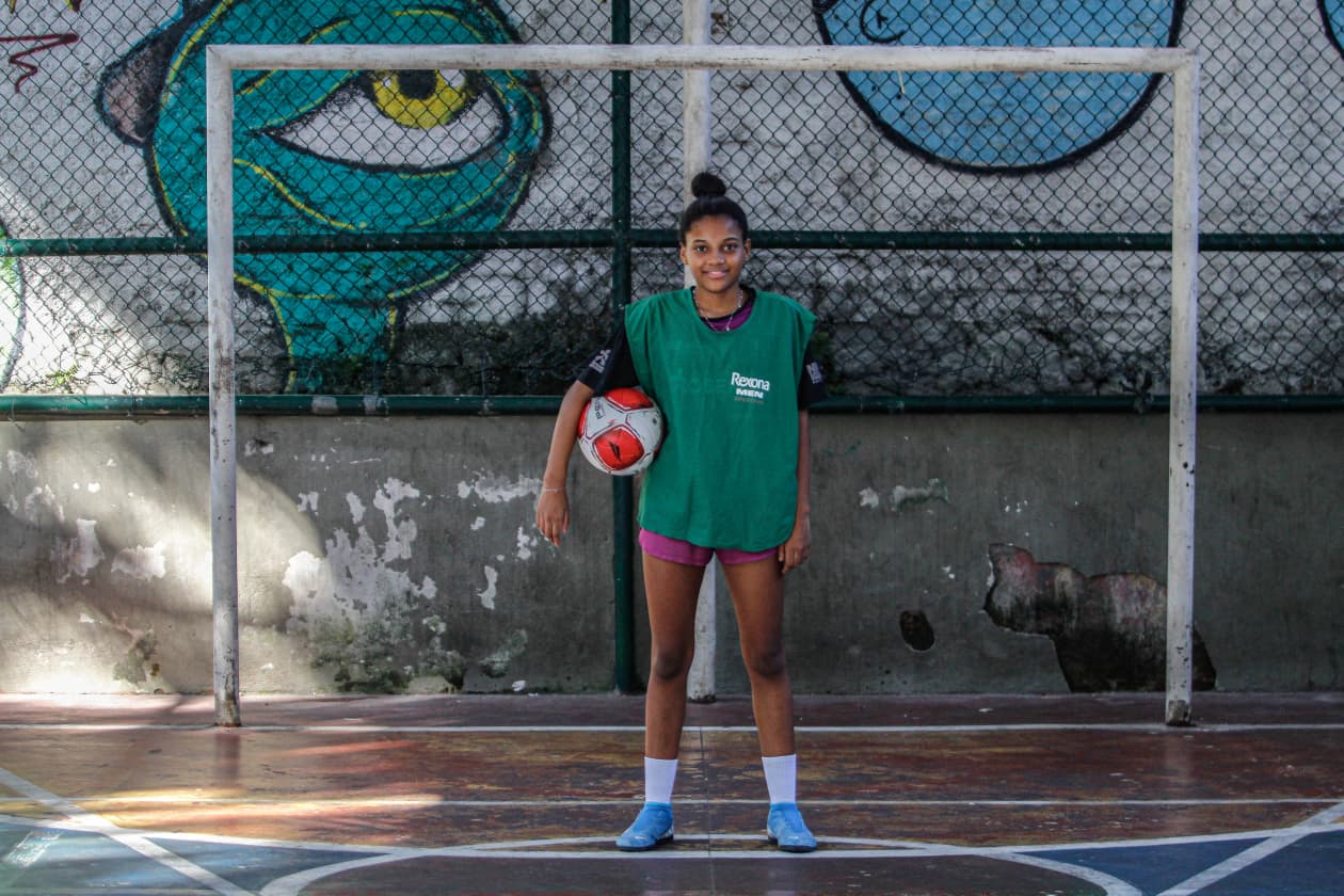 Essa imagem mostra uma jovem jogadora de futebol em pé no centro de um gol, segurando a bola com confiança. Ao fundo, um muro com grafites coloridos e marcas do tempo, transmitindo o contraste entre sonho e realidade no cenário urbano. Foto: Uendell Vinicius