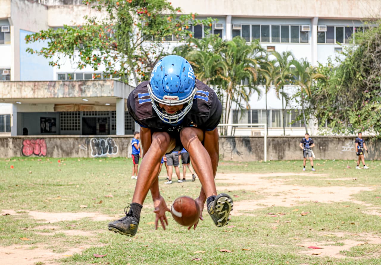 Um jovem jogador de futebol americano é capturado em pleno salto, prestes a agarrar a bola, transmitindo energia e intensidade. Ao fundo, companheiros de equipe assistem, compondo uma cena vibrante de treino ao ar livre. Foto: Uendell Vinicius